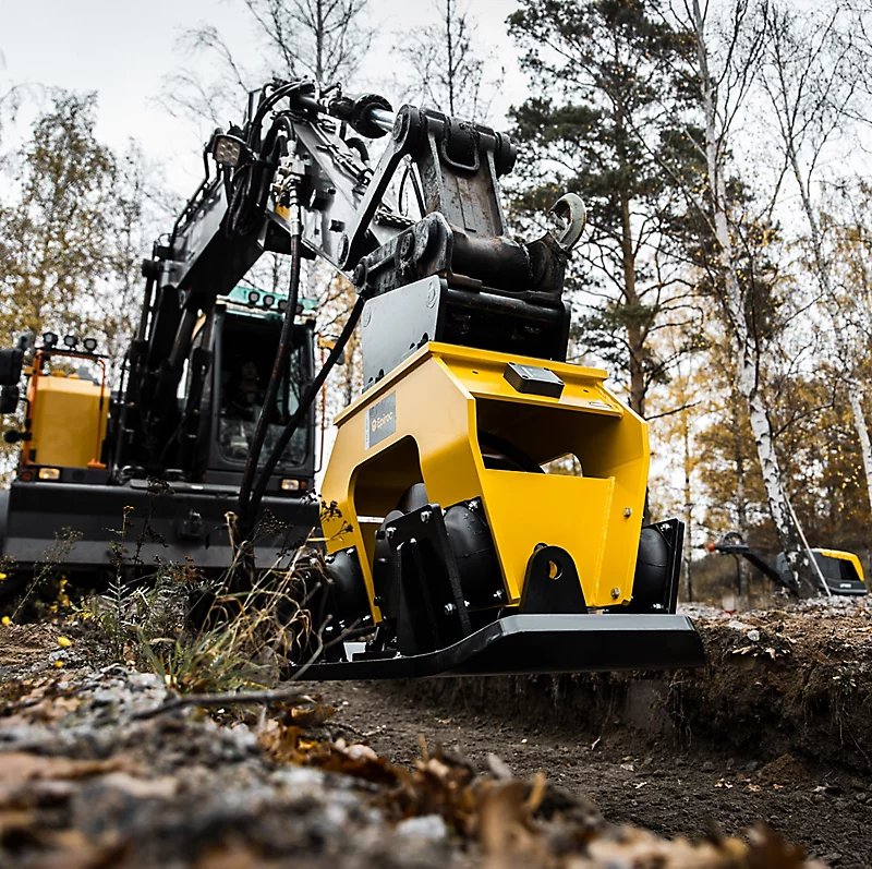 Excavator with a yellow attachment working on a construction site in a forested area.