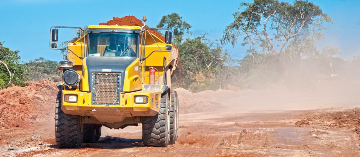 Yellow dump truck transporting soil on a construction site, creating dust clouds on a sunny day.