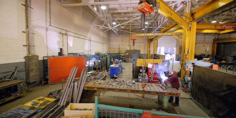A large industrial workshop with metal beams, tools, and machinery, where a worker in protective gear is welding at a table. Overhead cranes and various workstations, including OSC Equipment Rental Buffalo NY, are visible throughout the spacious, well-lit area.